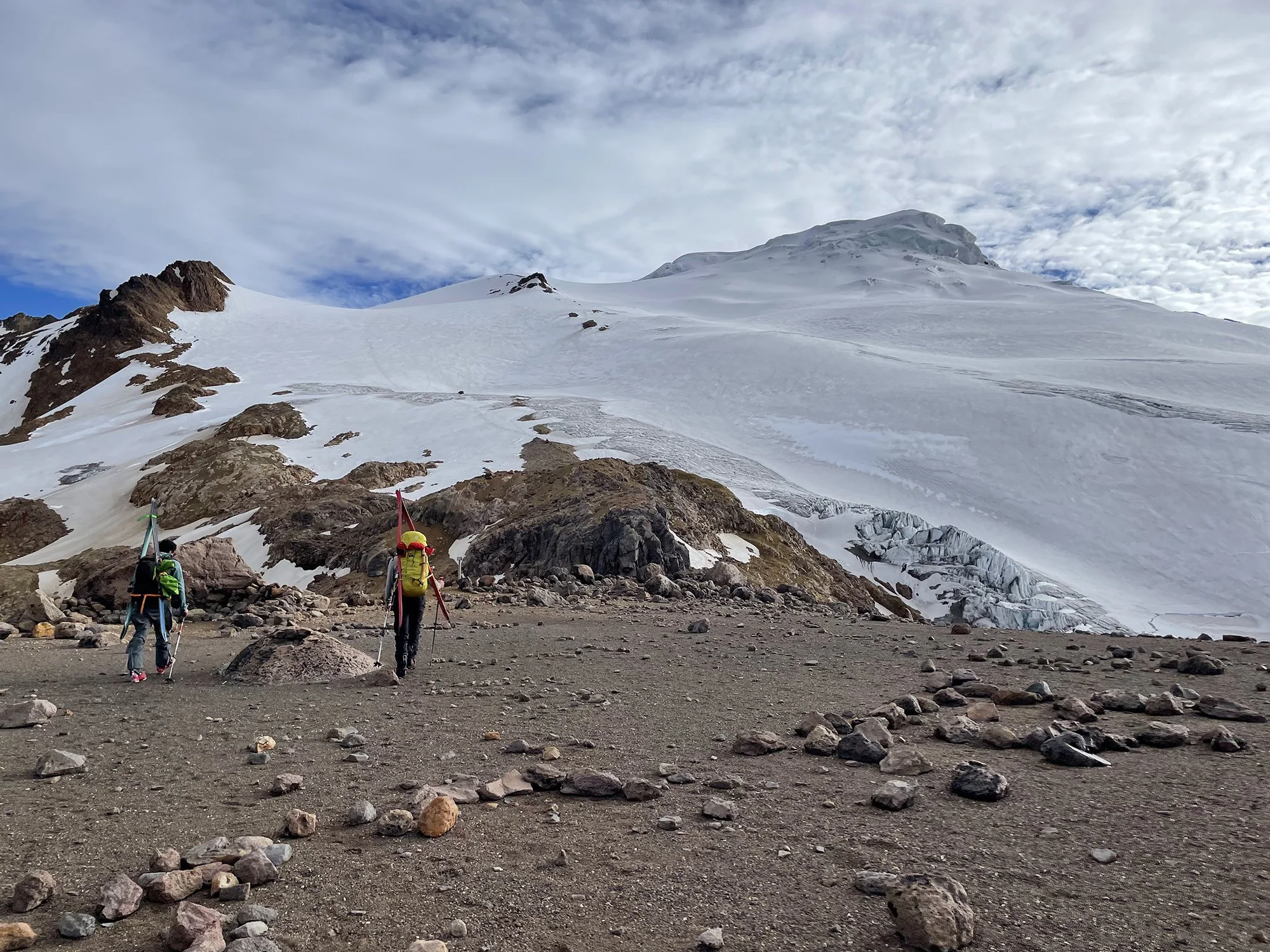 Three hikers walking on rocky terrain towards a snowy mountain with glaciers and a cloudy sky in the background.