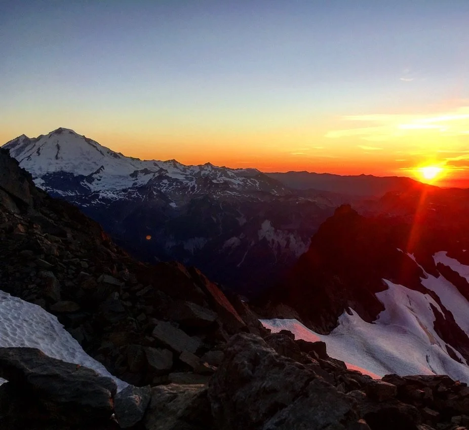 Mountain landscape at sunset with snow-covered peaks and rugged rocks in the foreground.