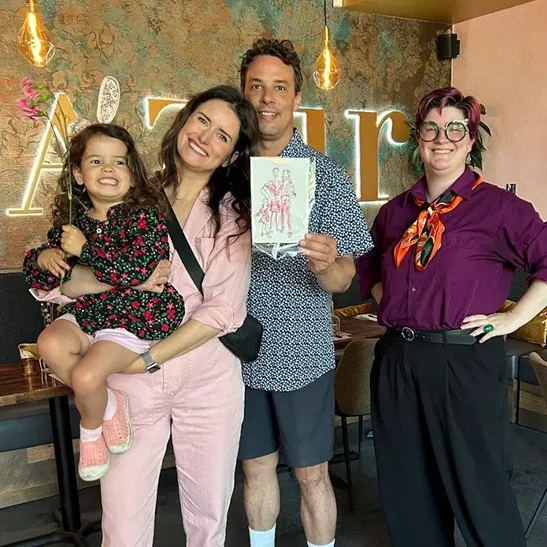 Brooke & Chris hold up an illustration of them created by Vignette Du Vin while holding their daughter in their arms and posing alongside the illustrator Spencer 