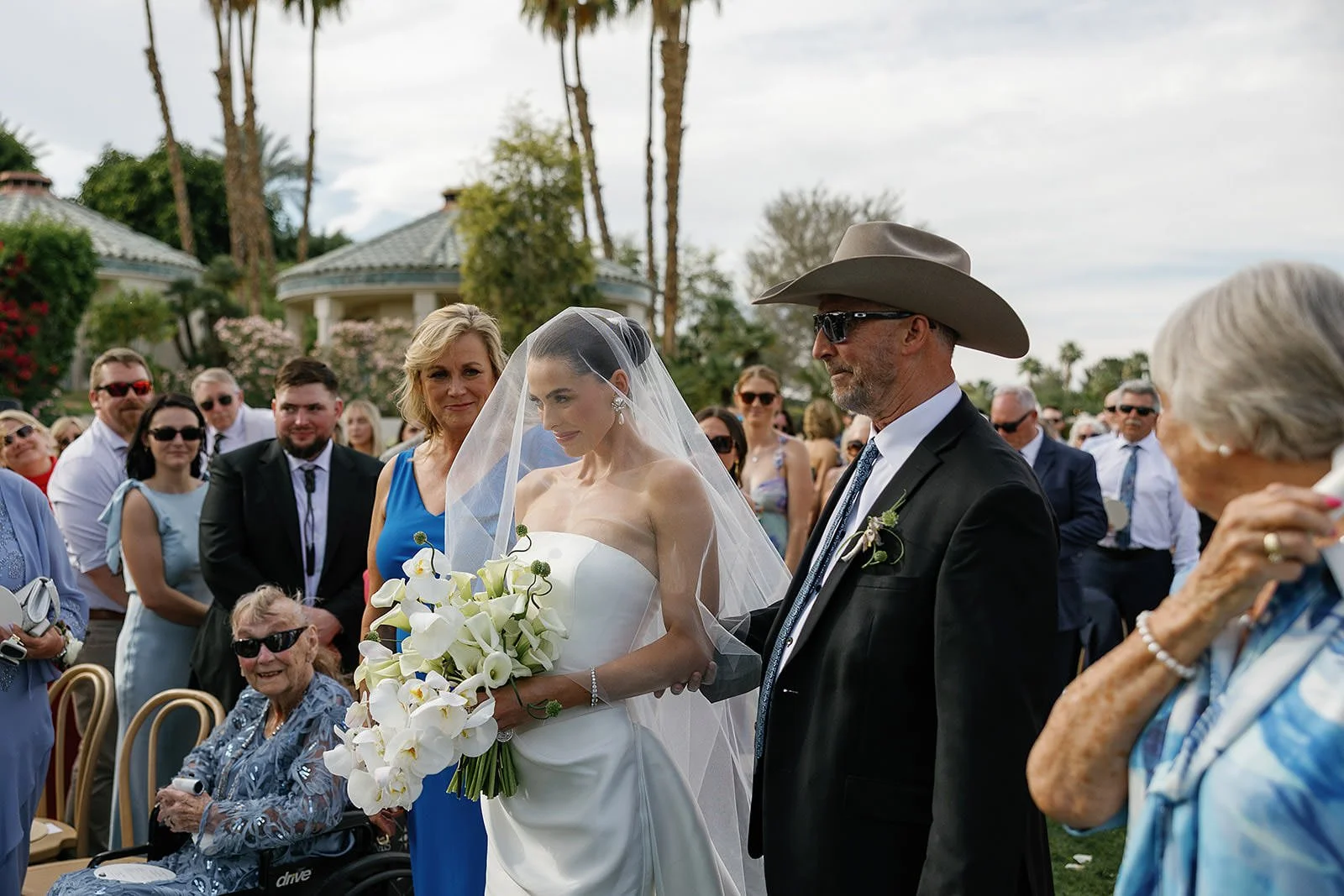 The bride and her parents walking down the aisle at a Merv Griffin Estate wedding ceremony with outdoor green garden party decor designed by COJ Events