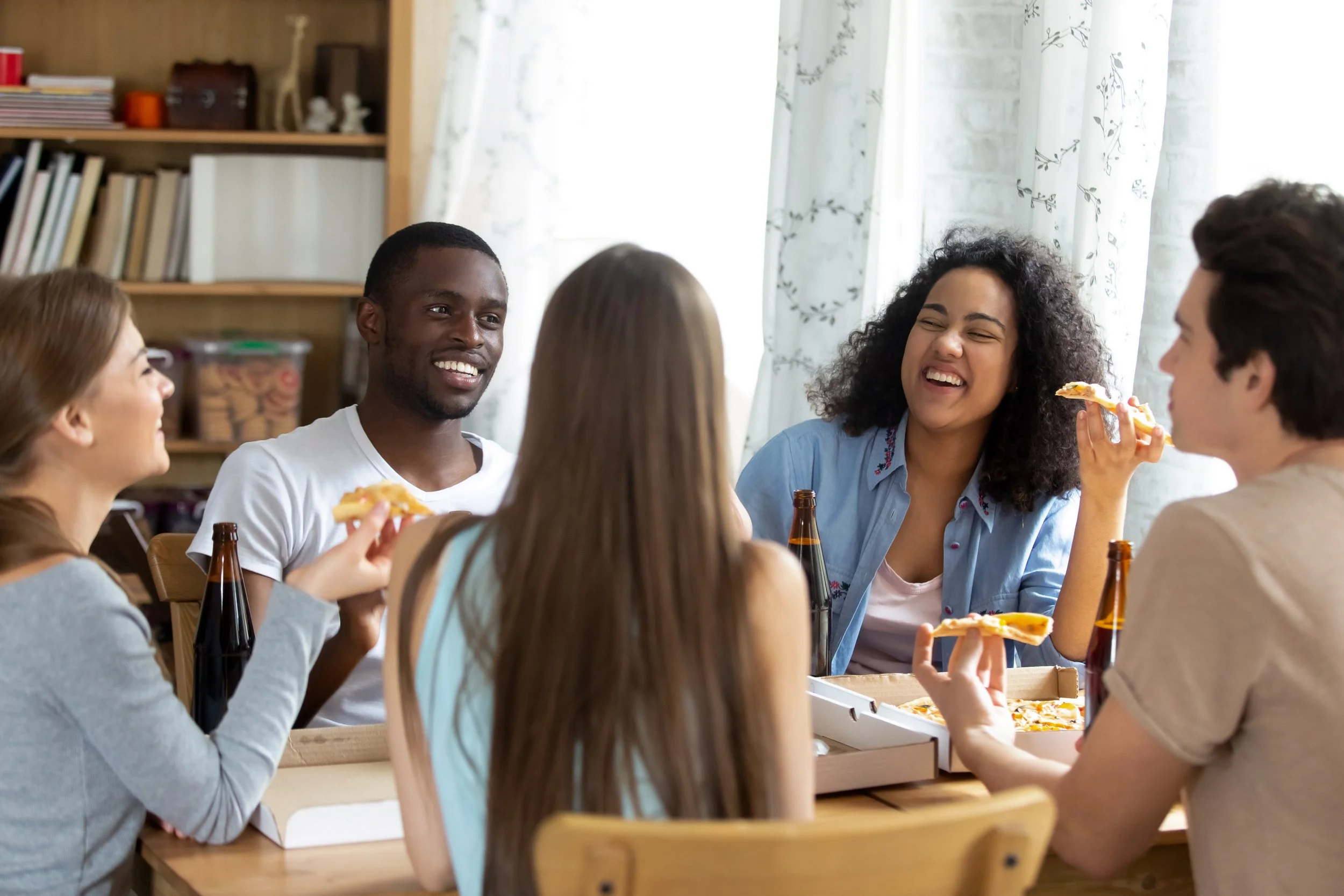 Group of five young adults, two women and three men, sitting at a wooden table enjoying pizza and drinks, smiling and laughing indoors.