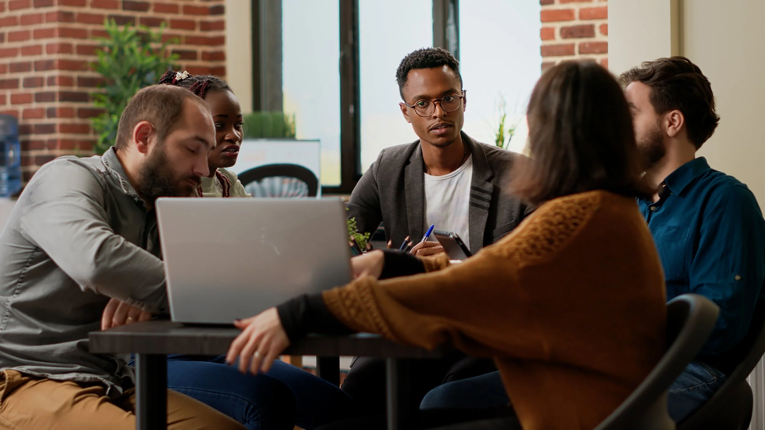Five people sitting around a table in a meeting room, engaged in discussion with laptops and notebooks.