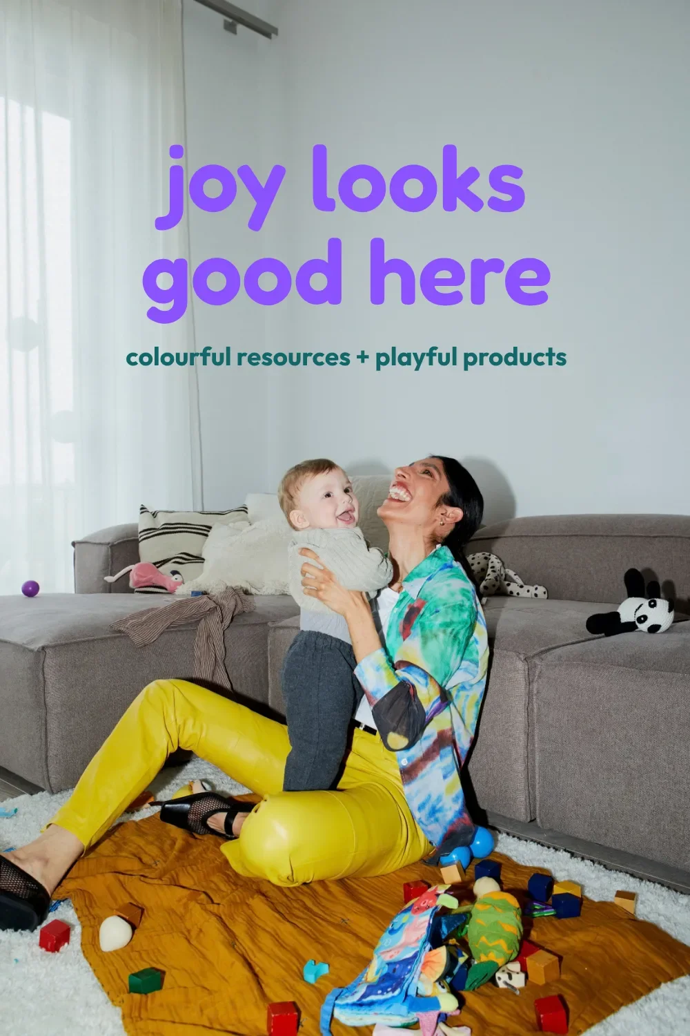 Mother laughing as she lifts her baby among scattered toys, with a purple joy looks good here headline