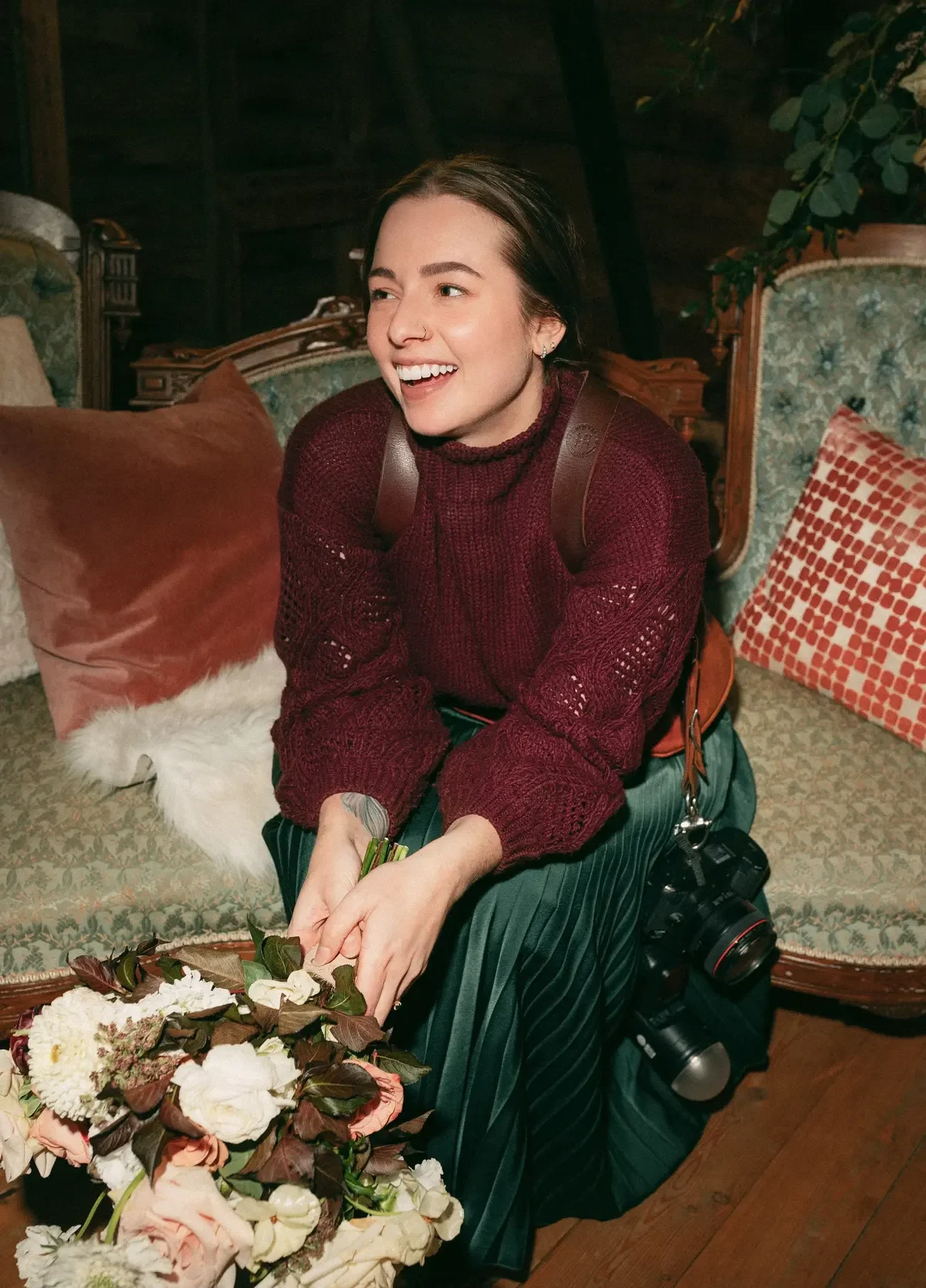 Laughing woman in a burgundy sweater holding a bouquet, with a camera at her hip, on a vintage sofa