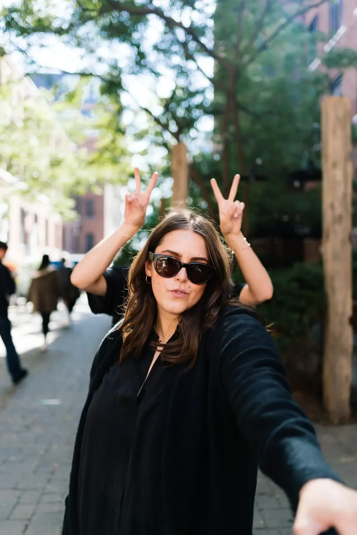 Woman in sunglasses taking a selfie as a friend makes bunny ears behind her on a tree-lined street