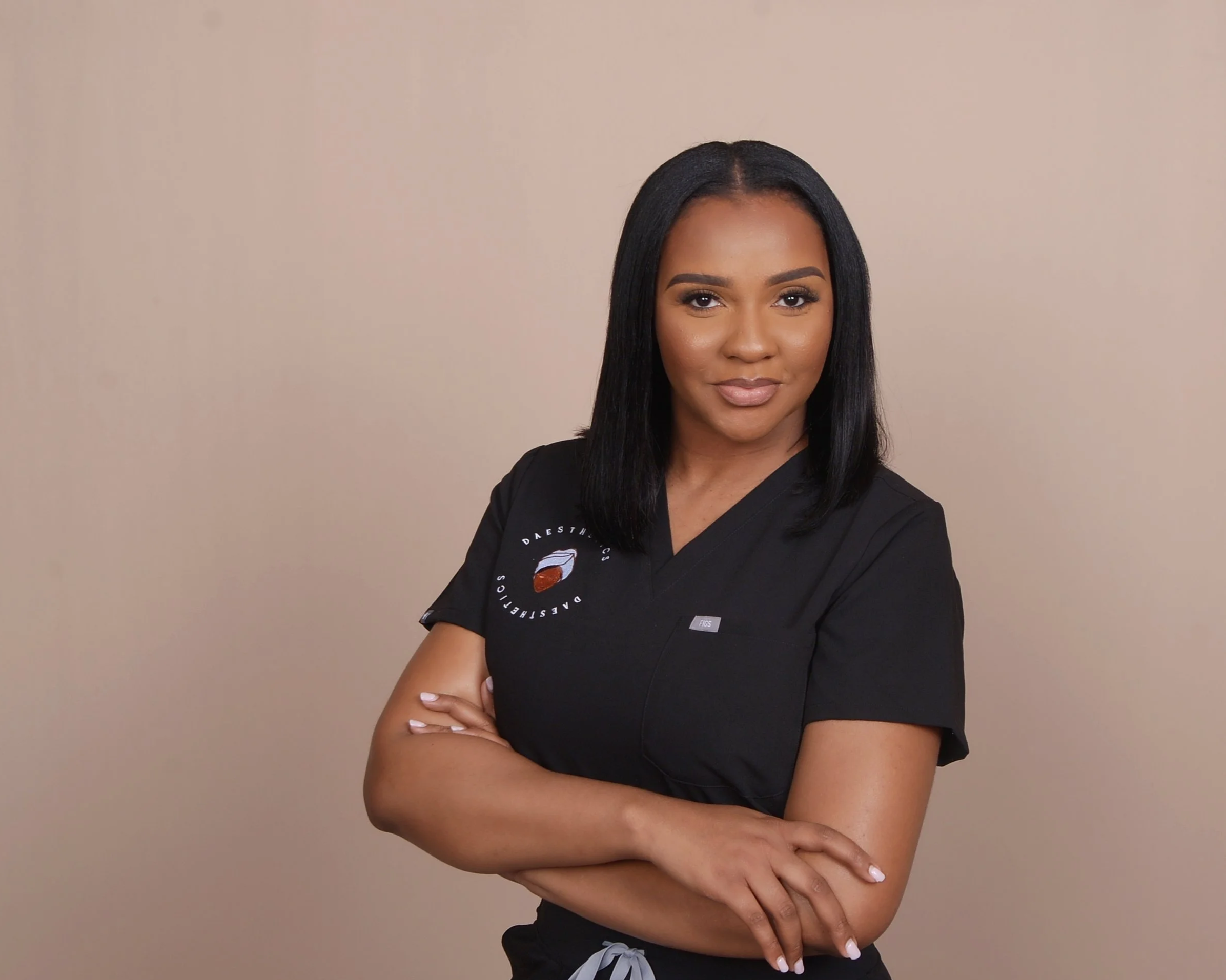 A confident woman with medium skin tone and straight black hair wearing a black medical uniform with her arms crossed, standing against a plain beige background.