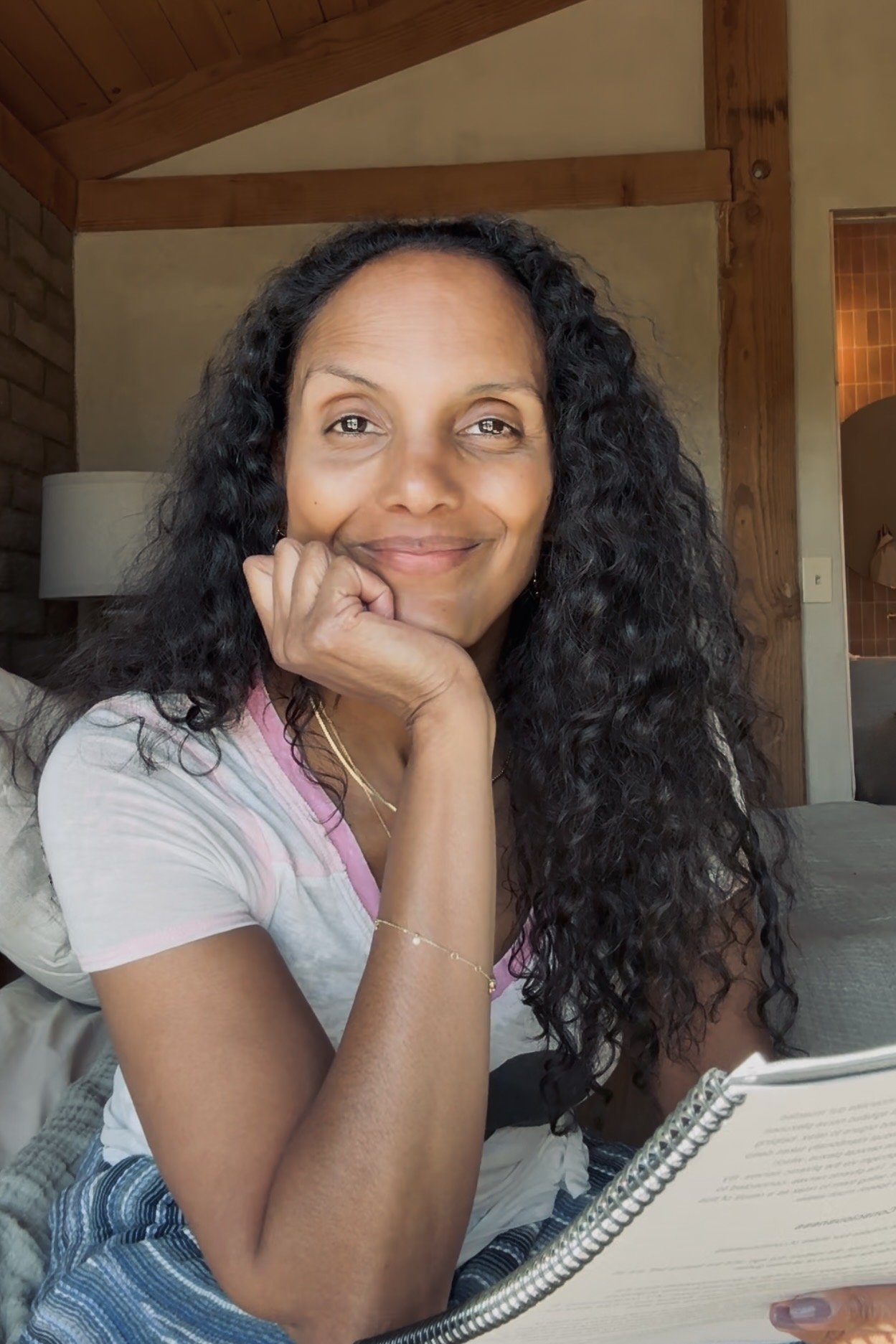 A woman with long, curly black hair smiling and resting her chin on her hand, holding a spiral notebook, sitting in a room with wood-paneled ceiling and beige walls.