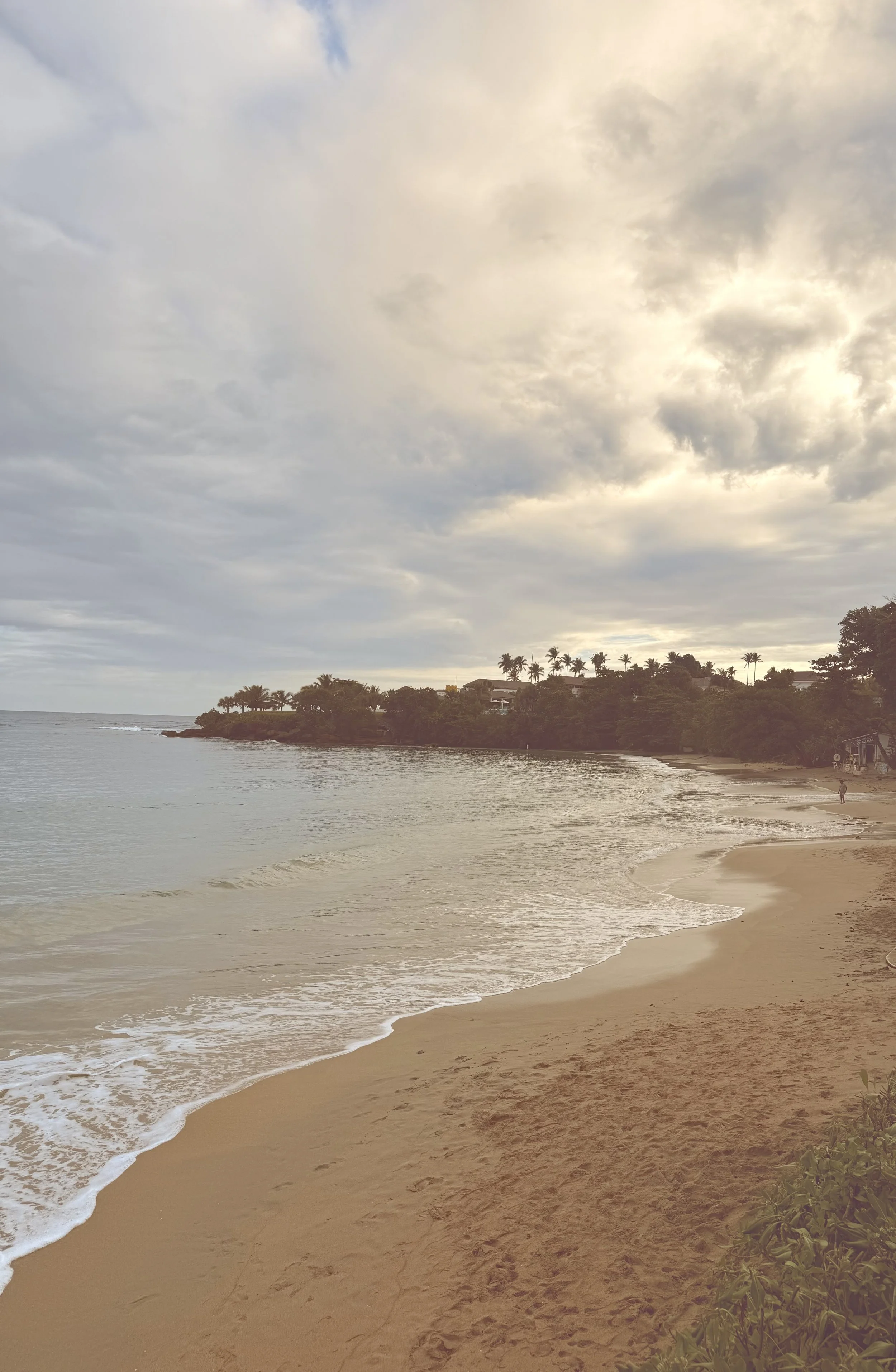A tranquil beach scene with sandy shore, gentle waves, and a cloudy sky. There are trees and buildings on a hill in the background, with a few people walking along the shoreline.