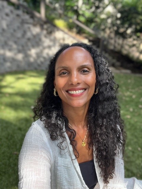 A woman with curly black hair smiling outdoors in a grassy area, wearing a light-colored jacket, earrings, and a gold necklace.