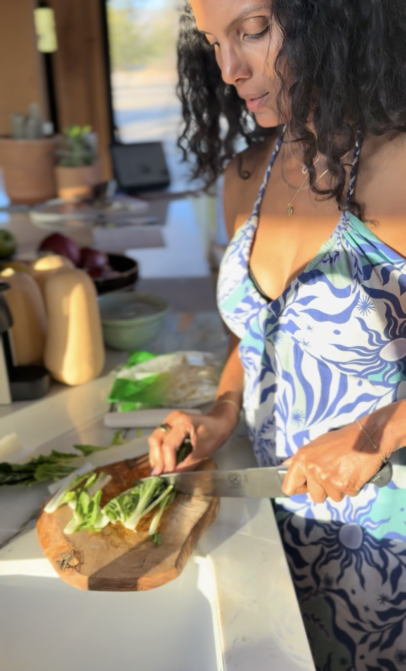 A woman with shoulder-length curly dark hair wearing a blue and white patterned dress chopping green vegetables on a wooden cutting board in a bright kitchen.