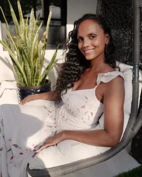A woman with dark, curly hair sitting on a round black outdoor chair, smiling, wearing a white dress with ruffled sleeves. There is a large potted plant with long green and yellow leaves behind her, and the setting appears to be a patio or outdoor space with sunlight.