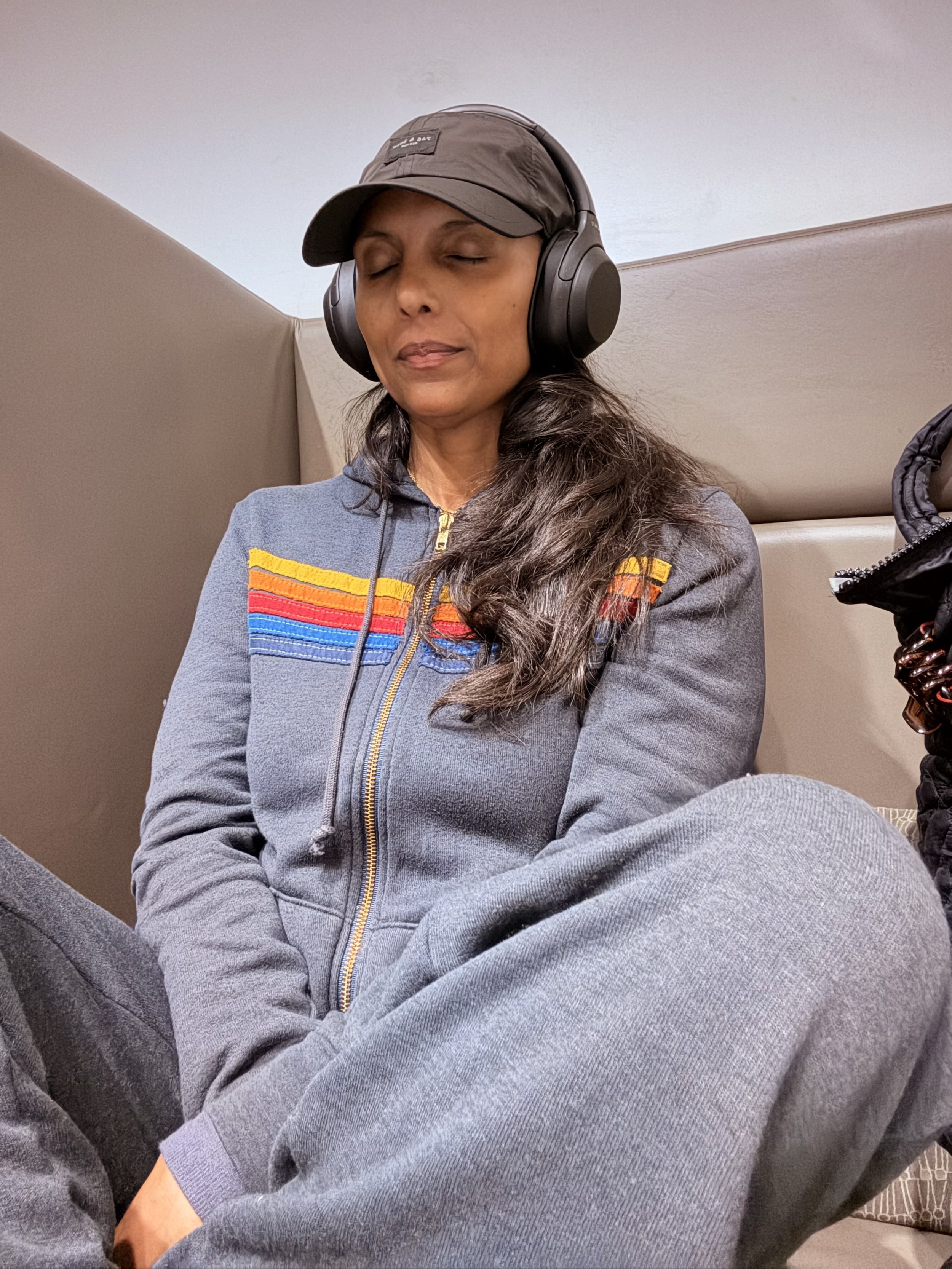 A woman sitting with her eyes closed, wearing headphones, a gray hoodie with colorful stripes, a black cap, and gray sweatpants.