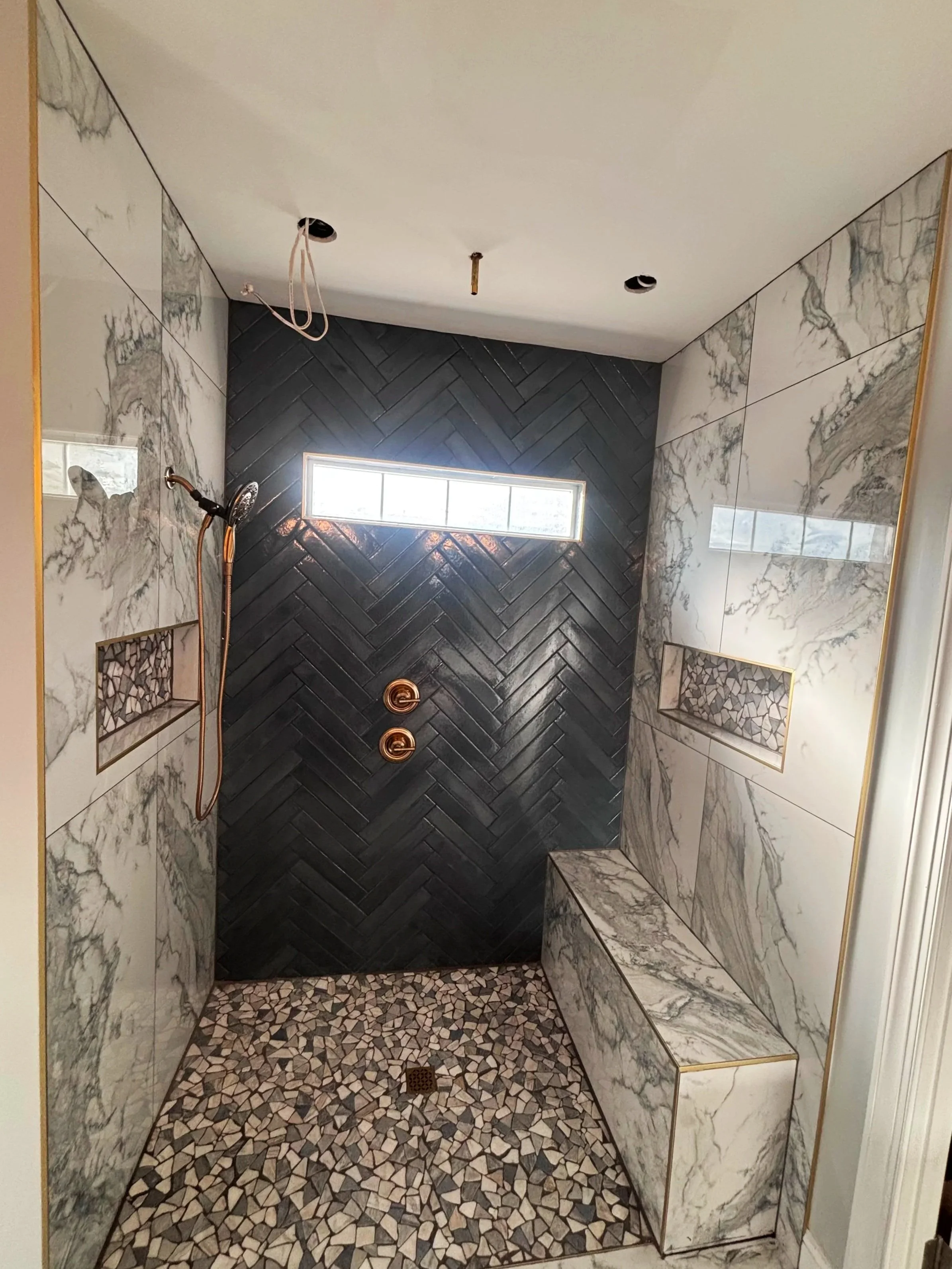 View of a modern walk-in shower with black herringbone tiles on the back wall, white marble tiles with gray veining on the side walls, pebble floor, built-in marble bench, and a small window for natural light.