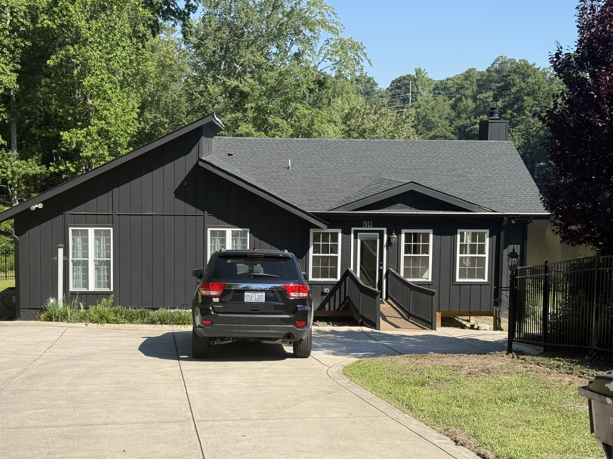 A gray house with a black roof and multiple windows, a black SUV parked in front, a small garden, and lush green trees surrounding the house on a clear, sunny day.