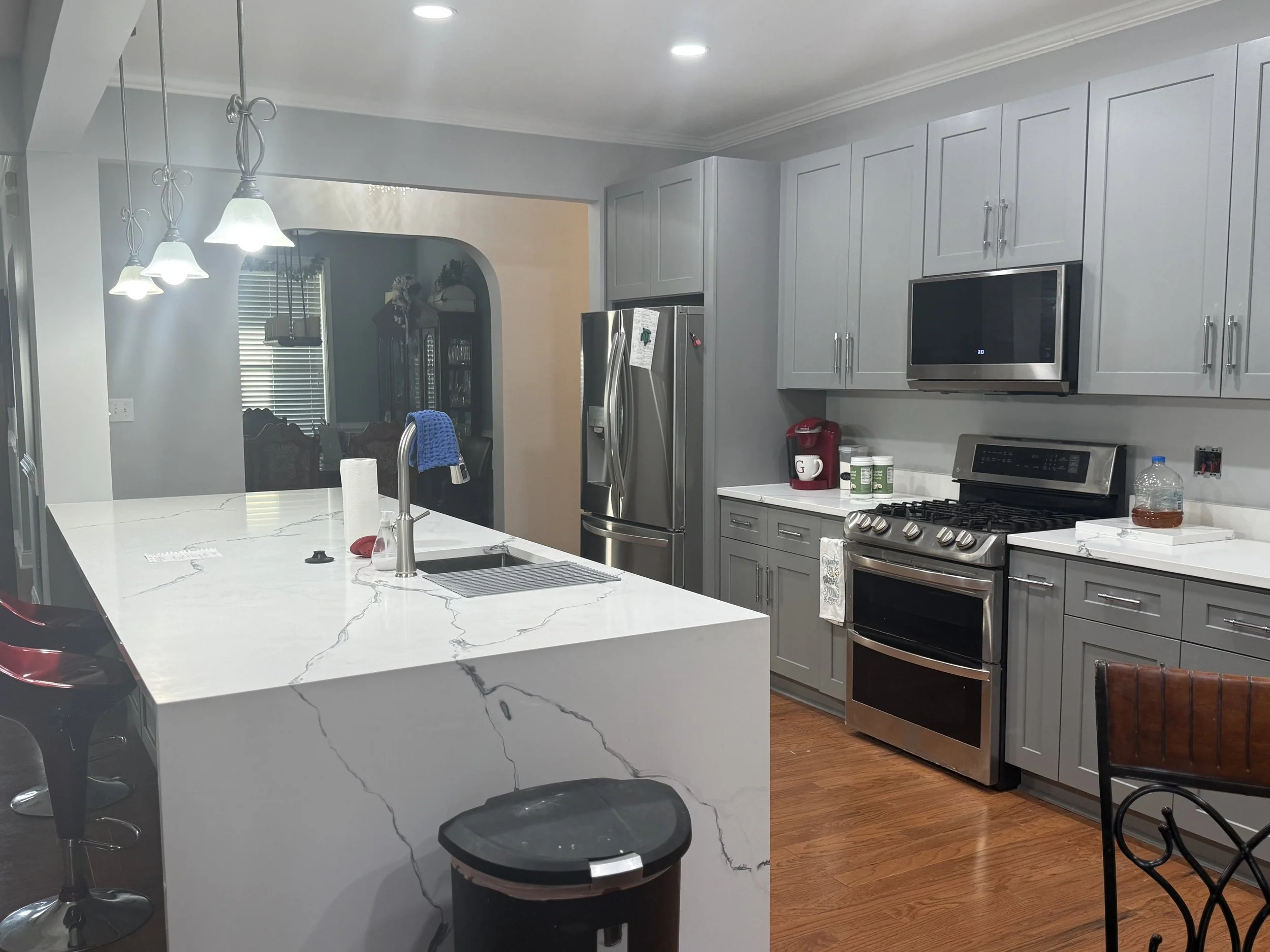 A modern kitchen with a white marble island, stainless steel appliances, and light gray cabinets. There are pendant lights above the island and a view into a dining area with large windows and blinds.
