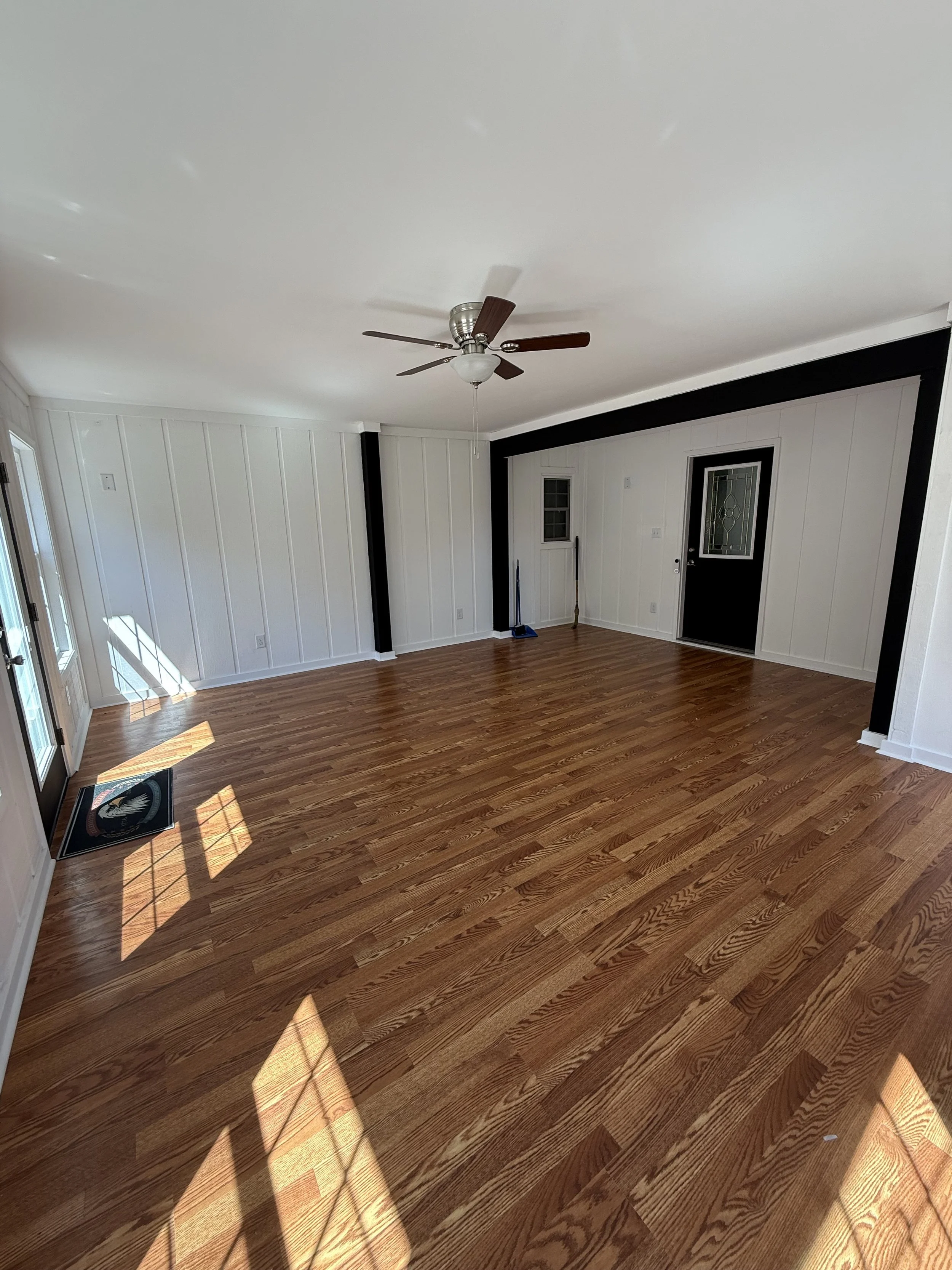 Empty living room with hardwood floors, white paneled walls with black accents, ceiling fan, and sunlight coming through glass door and windows.