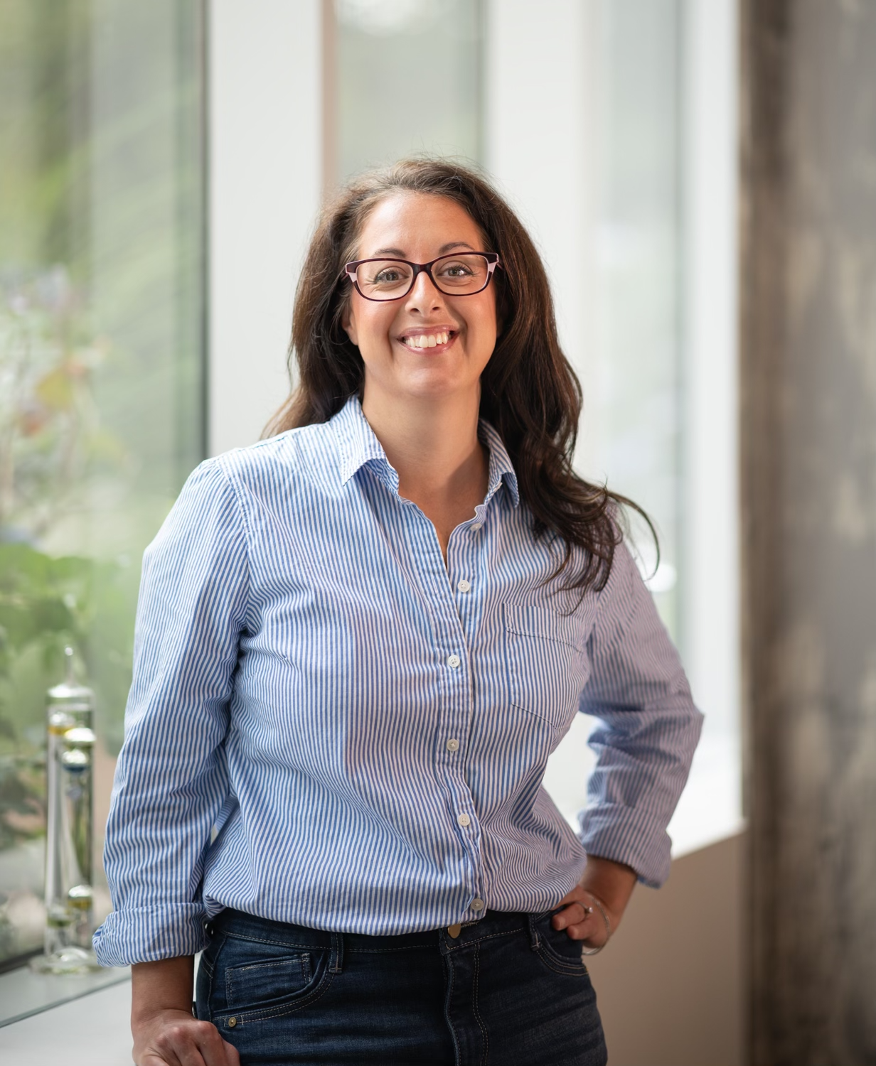 A woman with dark brown hair, glasses, and a striped blue button-up shirt standing indoors near a large window with plants outside, smiling confidently with one hand on her hip.