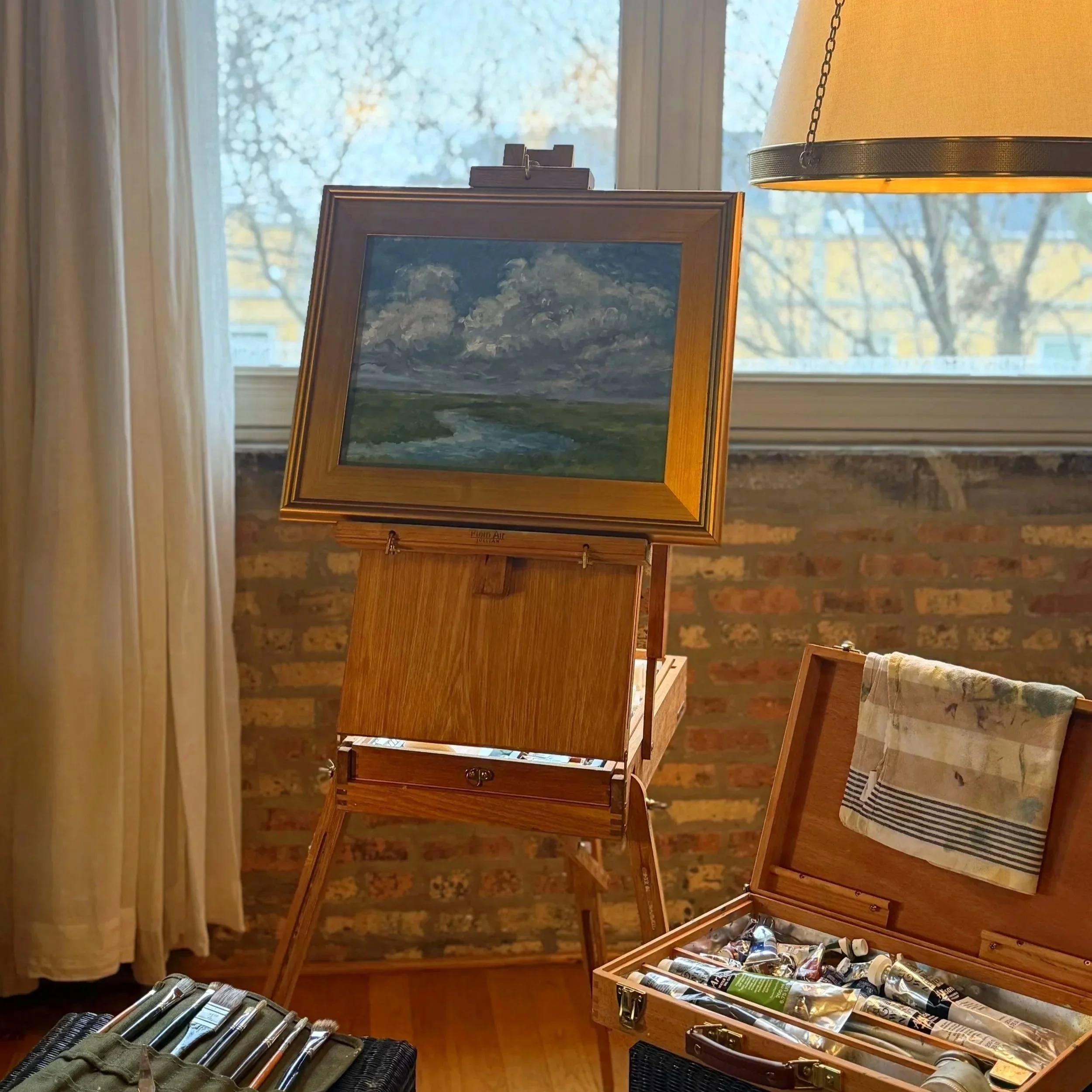 An art studio with a landscape painting of cloudy sky, river, and green fields, on an easel near a window.