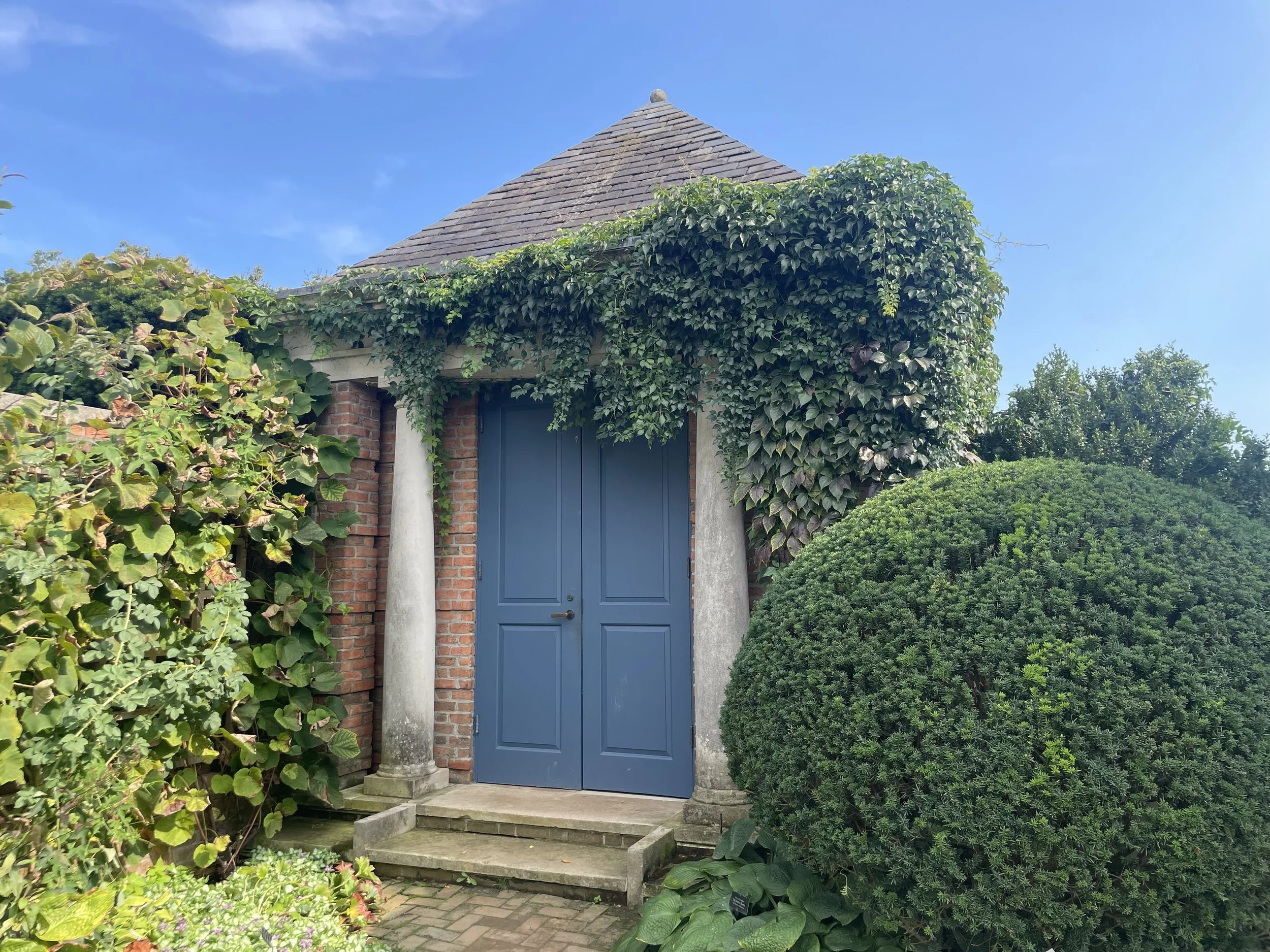 A small brick building with a blue door, surrounded by greenery and climbing plants, under a clear blue sky.