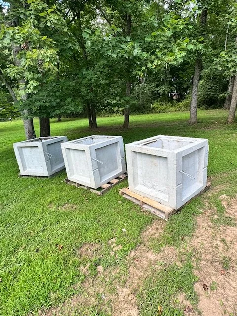 Three large white concrete or stone planters on wooden pallets in a grassy outdoor area with trees in the background.