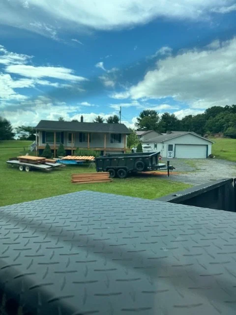 View of a backyard with a house, a garage, a black trailer, and stacks of wood on the grass, under a partly cloudy sky.
