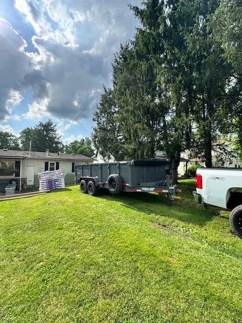 A yard with a trailer hitched to a white pickup truck, with tall trees and a cloudy sky in the background.