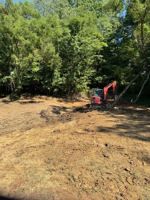 Construction site with an orange excavator and fresh dirt, surrounded by green trees.