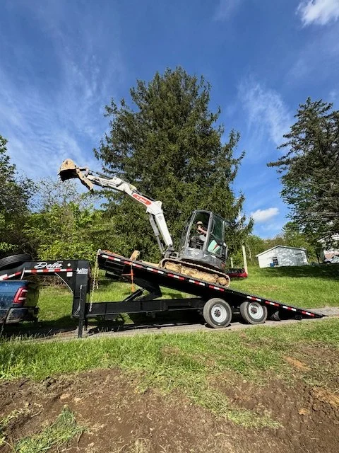 A small excavator on a flatbed trailer attached to a pickup truck, parked on a grassy area with trees and a house in the background.