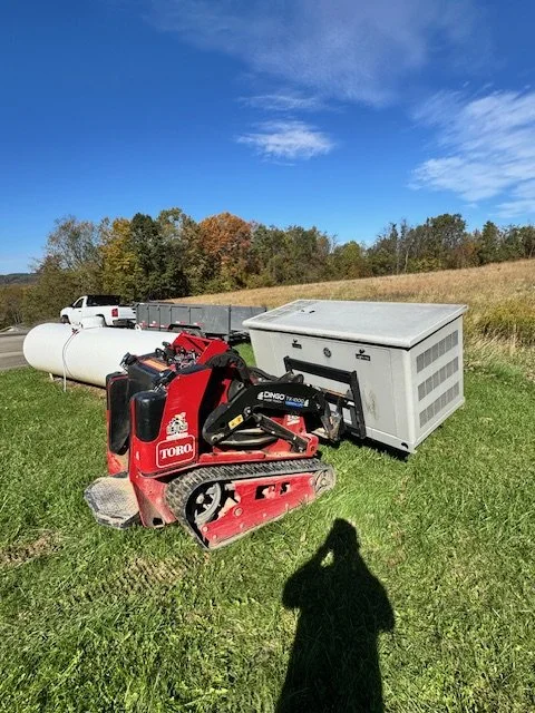 A small red Toro mini excavator next to a large industrial air conditioning unit on a grassy field with trees and a blue sky in the background.