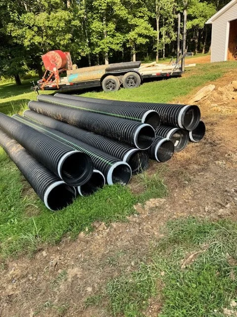 Stack of large black corrugated pipes on grass with a trailer and construction equipment in the background.