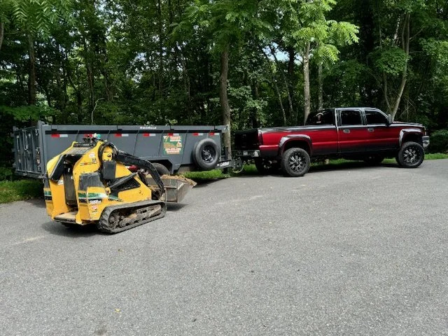 A small yellow construction skid steer loader parked on a paved surface next to a red pickup truck with black trim, connected to a black utility trailer, with a forest of green trees in the background.