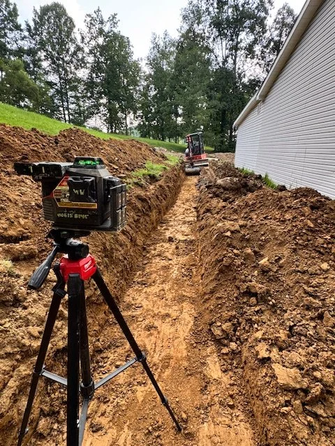 Construction site with a trench next to a house, with a laser level on a tripod in the foreground and a small excavator in the background.