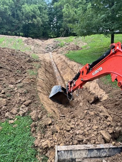 A Kubota mini excavator digging a trench in a grassy yard with trees in the background.