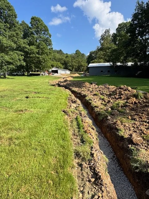 A large trench dug through a grassy yard, with gravel or small rocks at the bottom, near a farm or rural property with buildings and trees in the background under a partly cloudy sky.