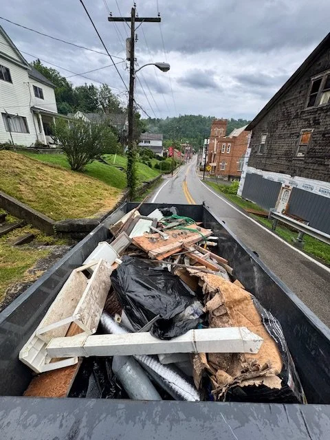 View of a dump truck filled with construction debris, including wood, metal, and insulation, on a residential street with houses, trees, and an overcast sky.