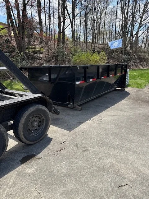 A large black dumpster attached to a flatbed truck parked on a paved road with trees and a flag in the background.