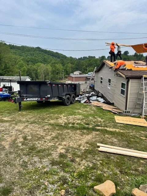 Workers on the roof of a damaged house repairing or removing the roof after a storm or disaster, with a trailer in the yard and debris scattered around.