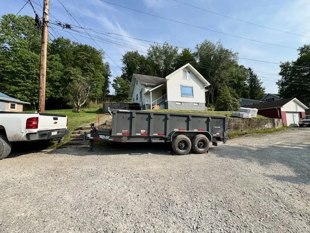 A black dump trailer hitched to a white pickup truck, parked on a gravel lot with a white house and trees in the background.