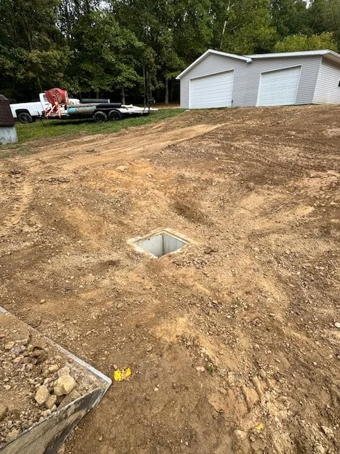 A yard under construction with a square utility access box exposed in the ground, a white garage, and a pickup truck in the background, surrounded by trees.