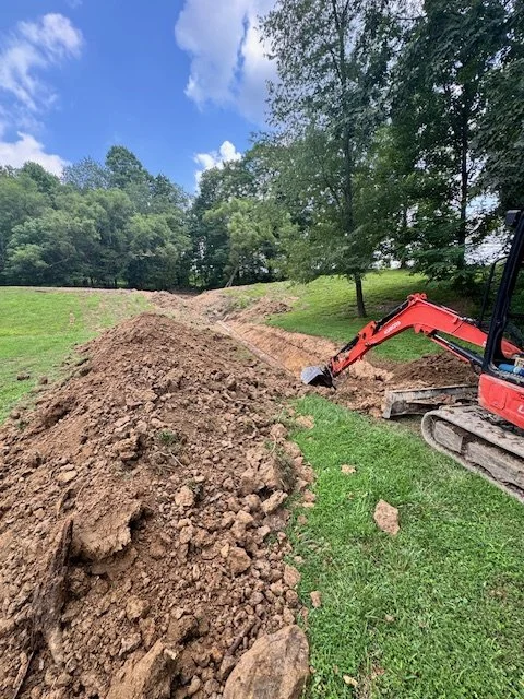 Construction site with an orange excavator digging a trench in a grassy area with trees and blue sky