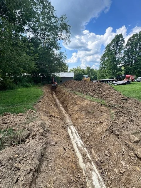 A construction site with a long trench and a pipe running through it, surrounded by dirt and grass, with trees and a blue sky with clouds in the background.