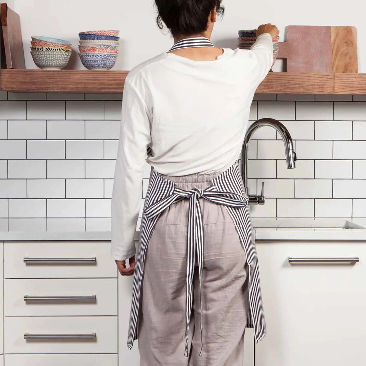 A person wearing a white shirt and a striped apron reaching for a bowl on a wooden shelf in a kitchen with white subway tile backsplash.