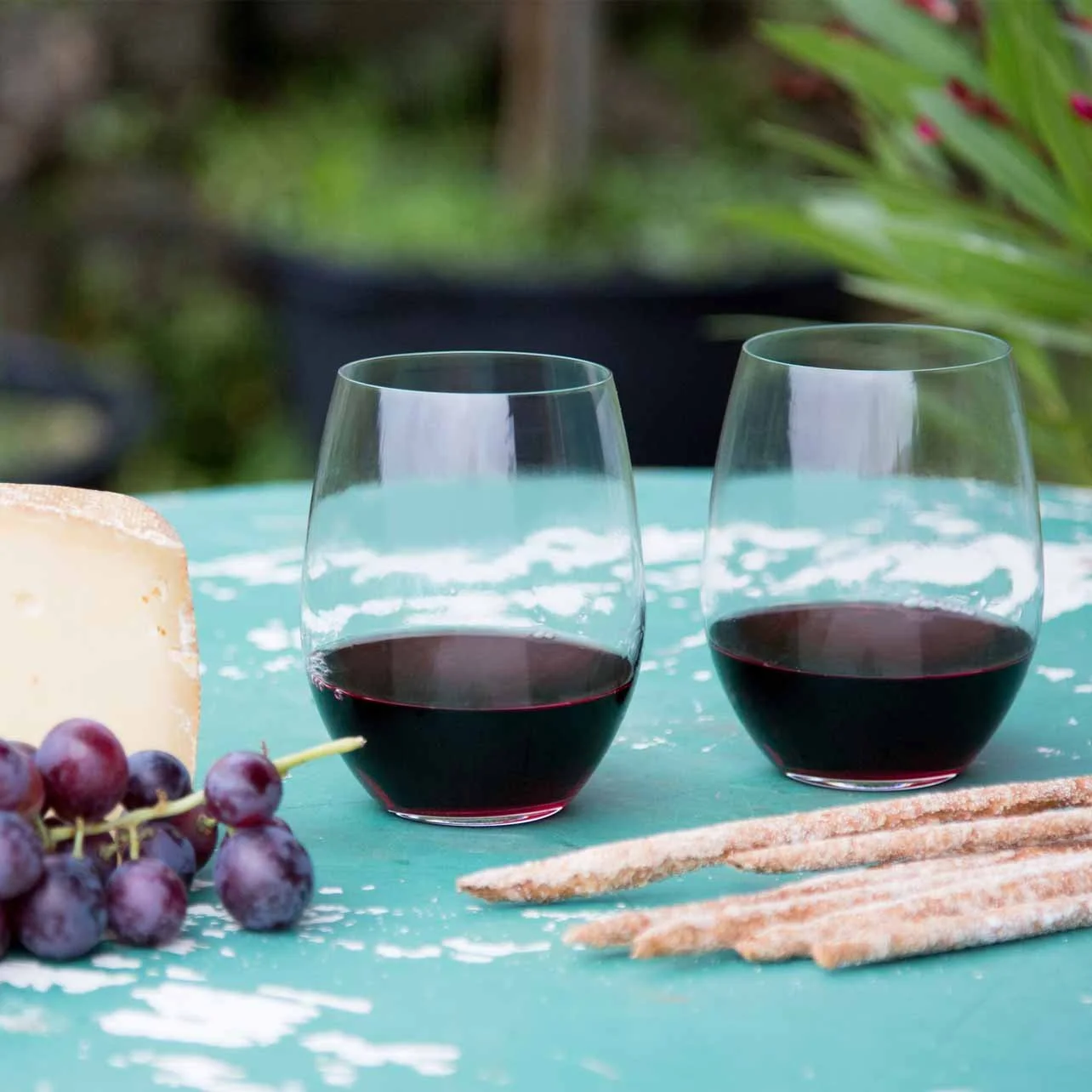 Two glasses of red wine on a turquoise table, with a wedge of cheese, a bunch of grapes, and breadsticks nearby, in an outdoor setting with green foliage background.