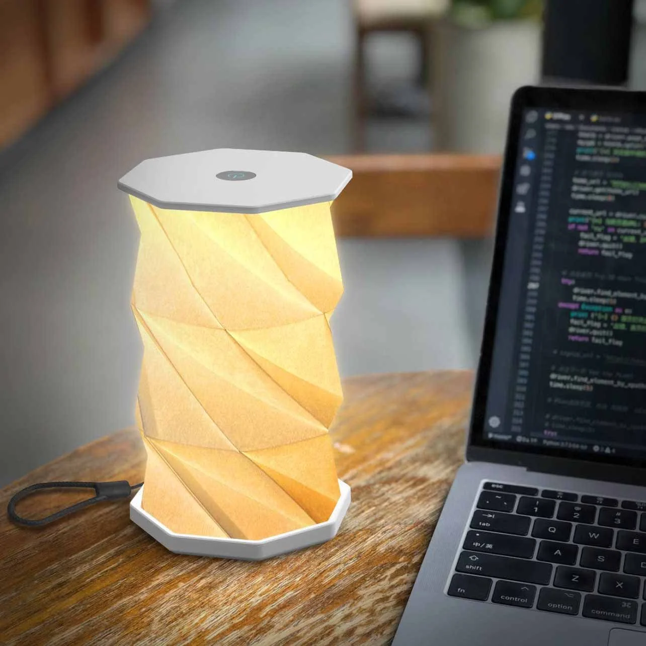 A modern table lamp with a spiral paper shade emitting warm light, placed on a wooden desk next to a laptop with coding on the screen, in a home or office setting.