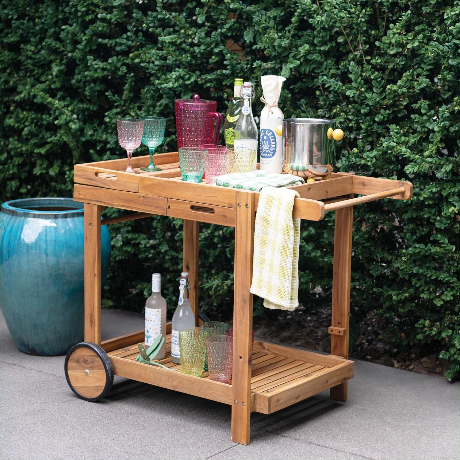 A wooden outdoor bar cart with colorful glassware, bottles, and a pot, positioned on a patio in front of green bushes.