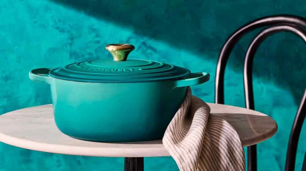 A teal ceramic Dutch oven on a white round table with a striped beige and white cloth draped over the edge, accompanied by a black metal chair with a curved backrest, set against a teal background.