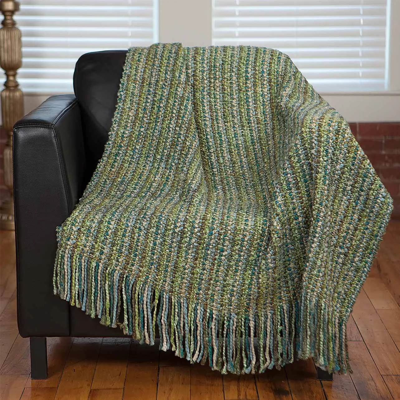 A colorful, textured blanket draped over the back and seat of a black leather sofa in a room with wooden floor and window blinds.