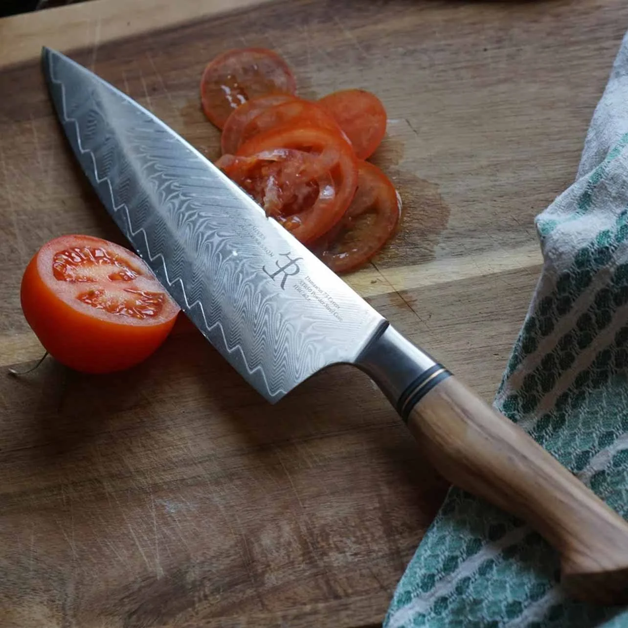 A wooden cutting board with a halved tomato, sliced tomatoes, a large kitchen knife, and a cloth towel.
