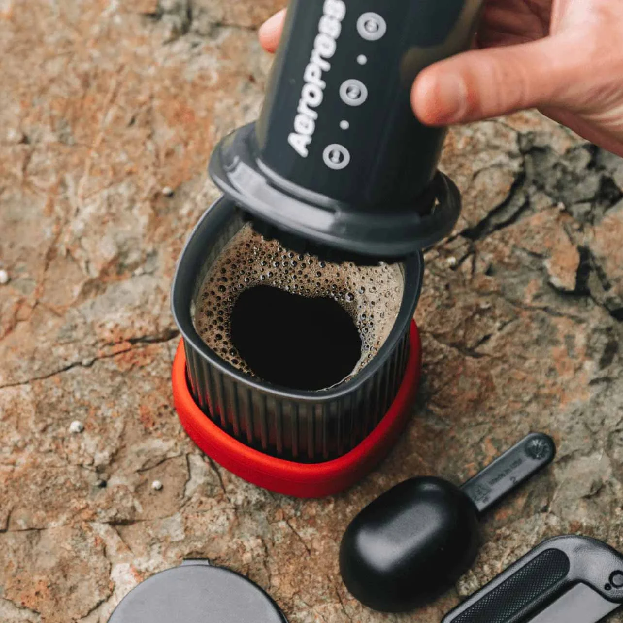 A hand pouring hot water from a AeroPress coffee maker into a cup on a rocky surface. Coffee bubbles are forming in the dark liquid, with a red base and a metallic filter inside the cup.