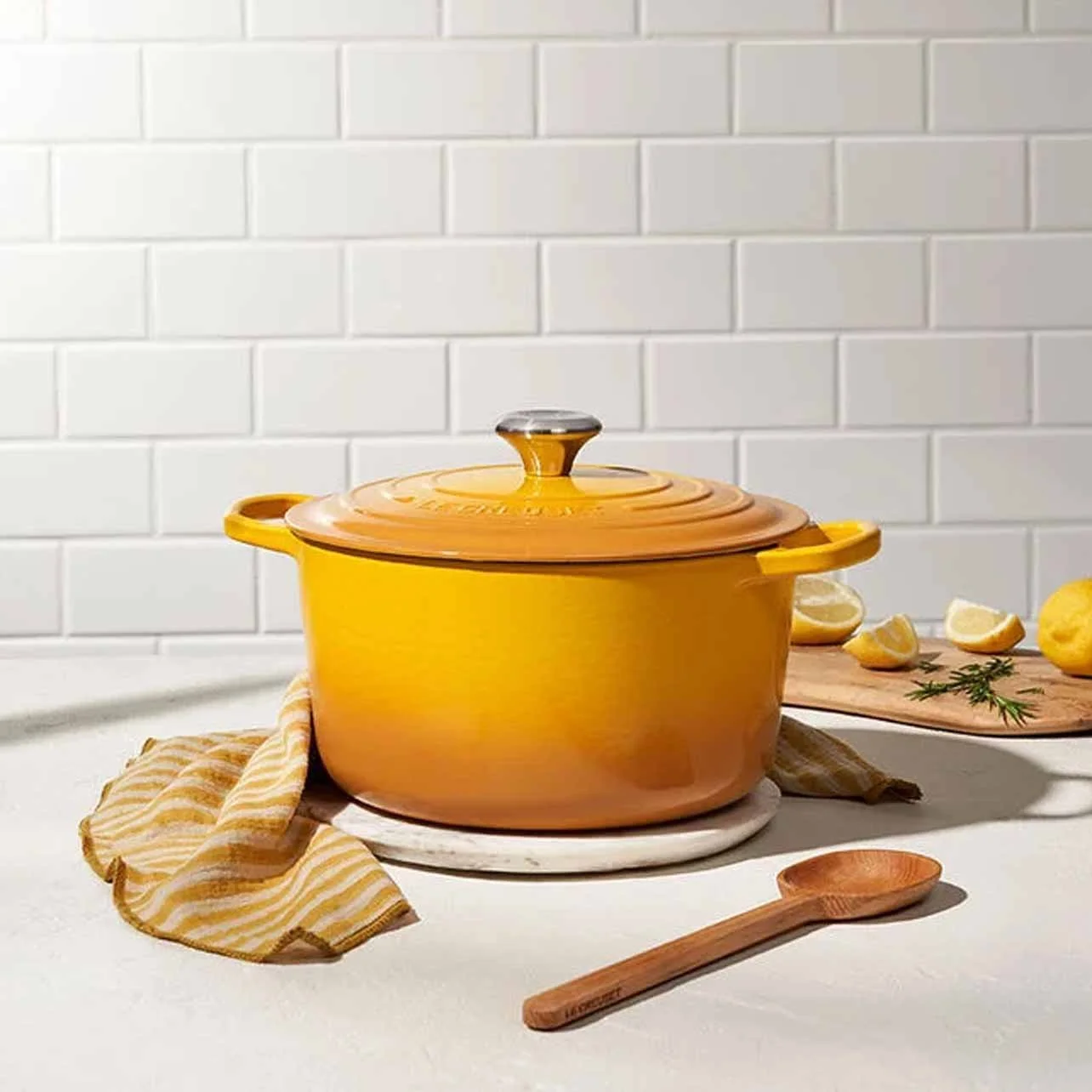 Yellow enameled cast iron pot with a glass lid in a kitchen setting, with lemons and sprigs of herbs on a wooden cutting board nearby, and a wooden spoon on the countertop.